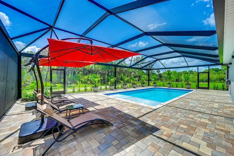 Pool in a screened lanai with red cantilever umbrellas and loungers on a paver deck